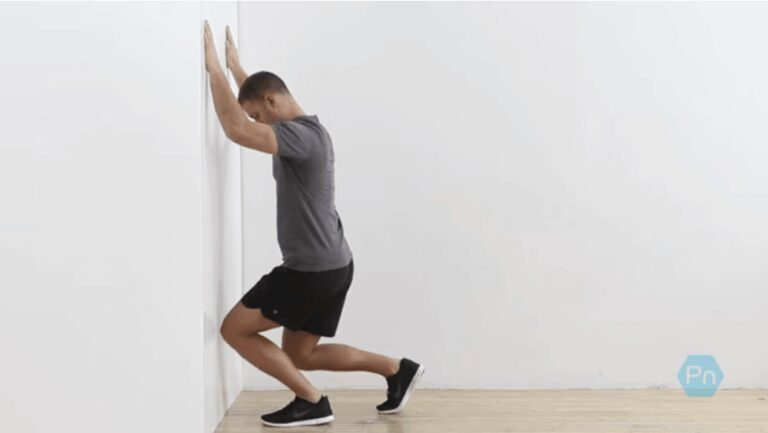 A man performing a standing wall ankle mobilization drill, pushing his knee towards the wall to stretch the calf and improve ankle dorsiflexion, which is essential for deep squats and running.